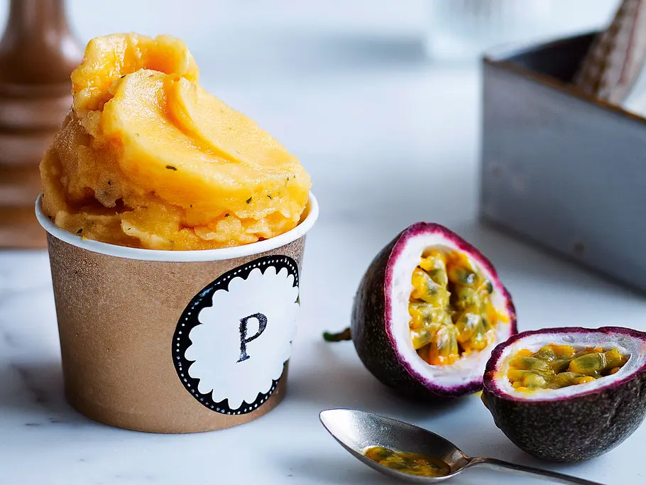 Passionsfruchtsorbet mit Minze Rezept Ein kleiner Eisbecher aus Pappe voll gefüllt mit gelben Passionsfruchtsorbet, daneben eine aufgeschnittene Passionsfrucht