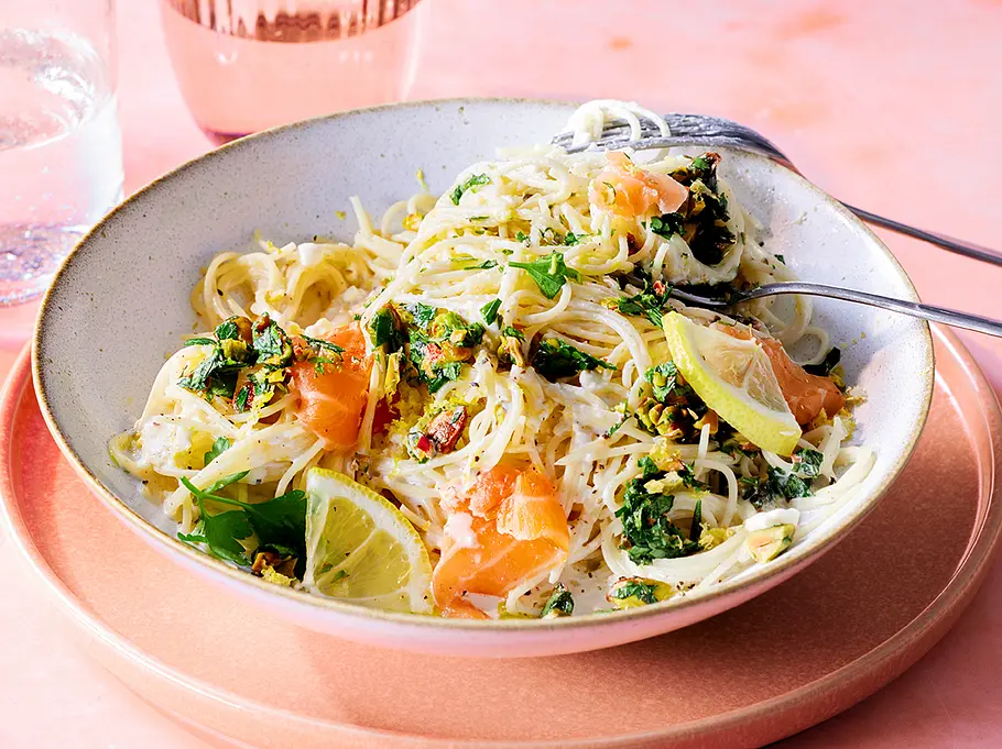 Spaghetti mit cremiger Frischkäsesoße, Räucherlachs, frischen Kräutern und Zitronenspalten auf einem Teller angerichtet.