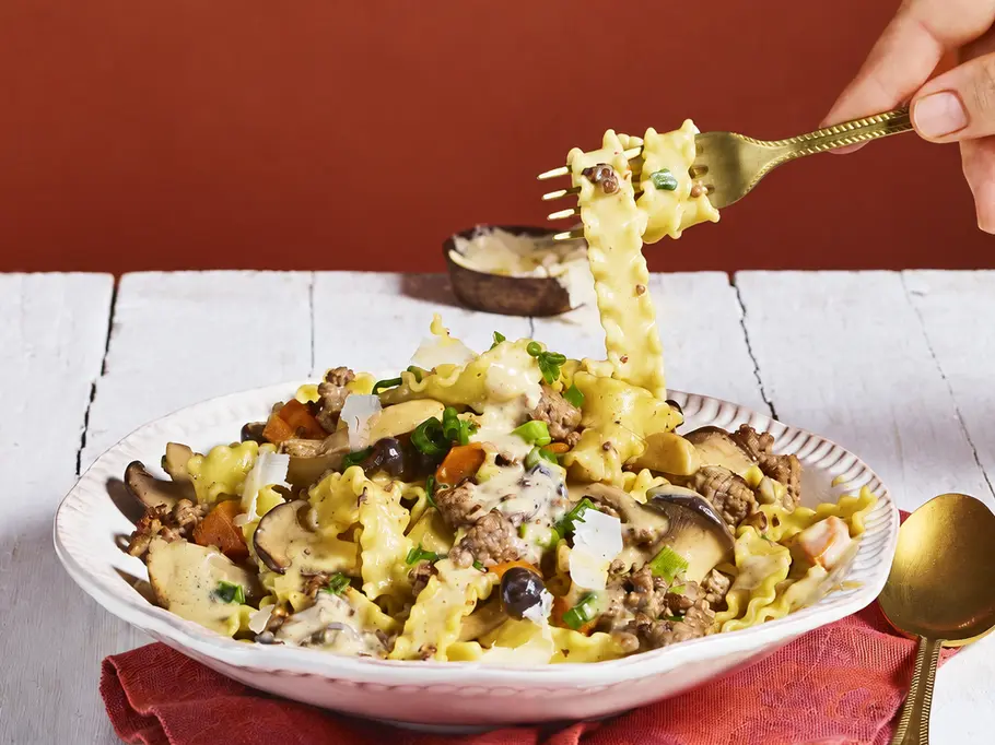 Pasta mit Jäger-Bolo Eine Hand hebt mit einer goldenen Gabel Bandnudeln vom Teller. Die Pasta ist mit Pilzen, Gemüse, Käse und cremiger Sauce angerichtet.