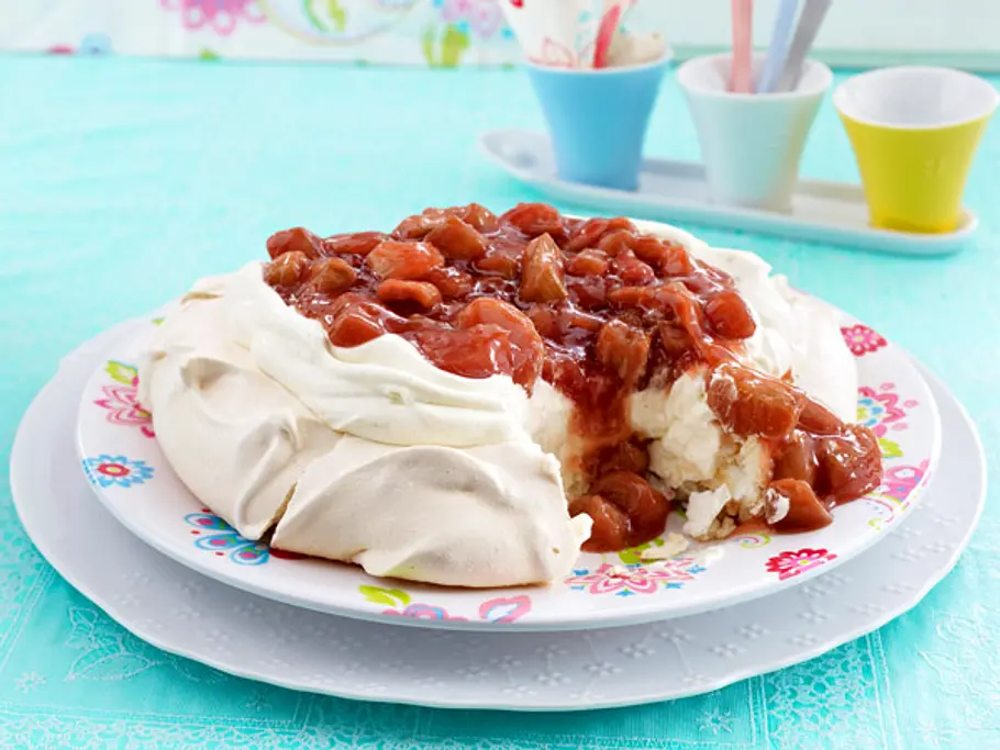 Eine Pavlova mit knusprigem Baiserboden, gefüllt mit Sahne und Rhabarberkompott, angerichtet auf einem bunten Teller, servierfertig auf einem hellblauen Tischtuch.