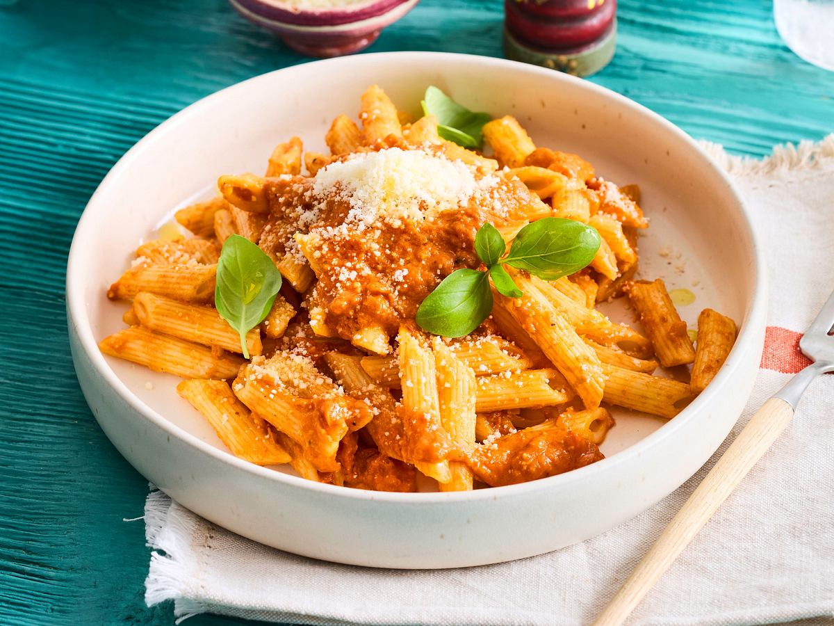Penne mit Tomatensauce, Basilikum und geriebenem Parmesan liegen in einer Schale auf einem hellen Tuch, daneben ein Besteck.