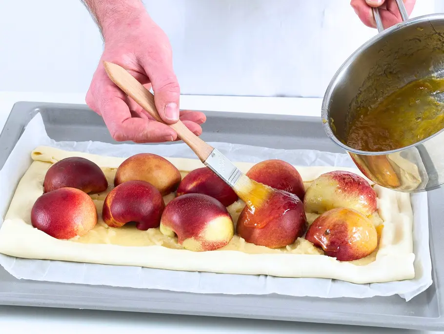 Blätterteig ausgerollt auf einem Backblech, darauf eine Marzipanmasse und halbierte Pfirsiche, die mit Aprikosenkonfitüre bestrichen werden.