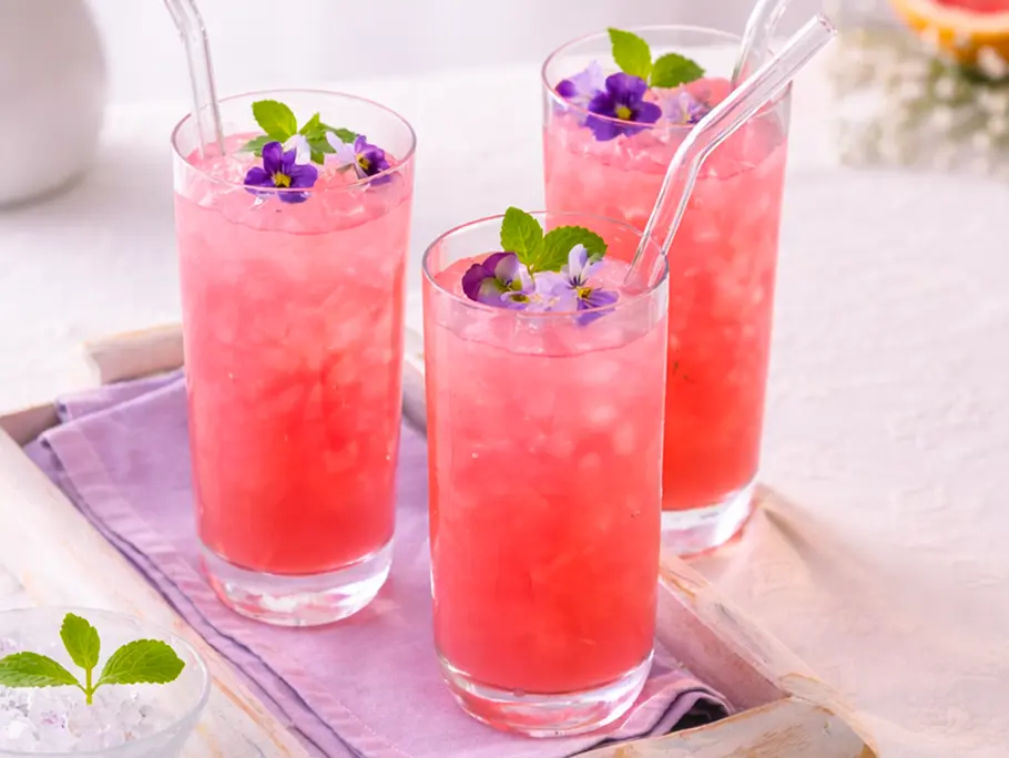 Pink Grapefruit Spritz Rezept 3 Longdrinkgläser mit rosafarbenem Inhalt stehen auf einem Holztablett mit lila Serviette, im Hintegrund sieht man Blümchen und eine Grapefruithälfte