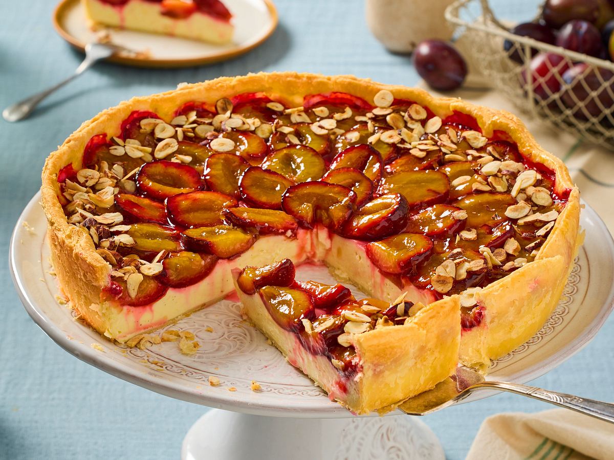 Pflaumen-Schmand-Kuchen auf einer Tortenplatte, angeschnitten. Ein Stück auf Tortenheber