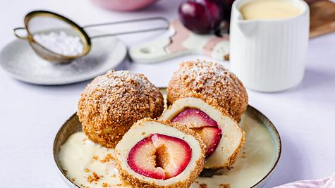 Drei Zwetschgenknödel sind auf einem Teller mit Vanillesoße serviert. Zu erkennen ist die knusprige Bröselkruste. Ein Pflaumenknödel ist halbiert, sodass man die Frucht erkennt. - Foto: ShowHeroes für LECKER @ Bauer Media Group