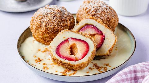 Drei Zwetschgenknödel sind auf einem Teller mit Vanillesoße serviert. Zu erkennen ist die knusprige Bröselkruste. Ein Pflaumenknödel ist halbiert, sodass man die Frucht erkennt. - Foto: ShowHeroes für LECKER @ Bauer Media Group