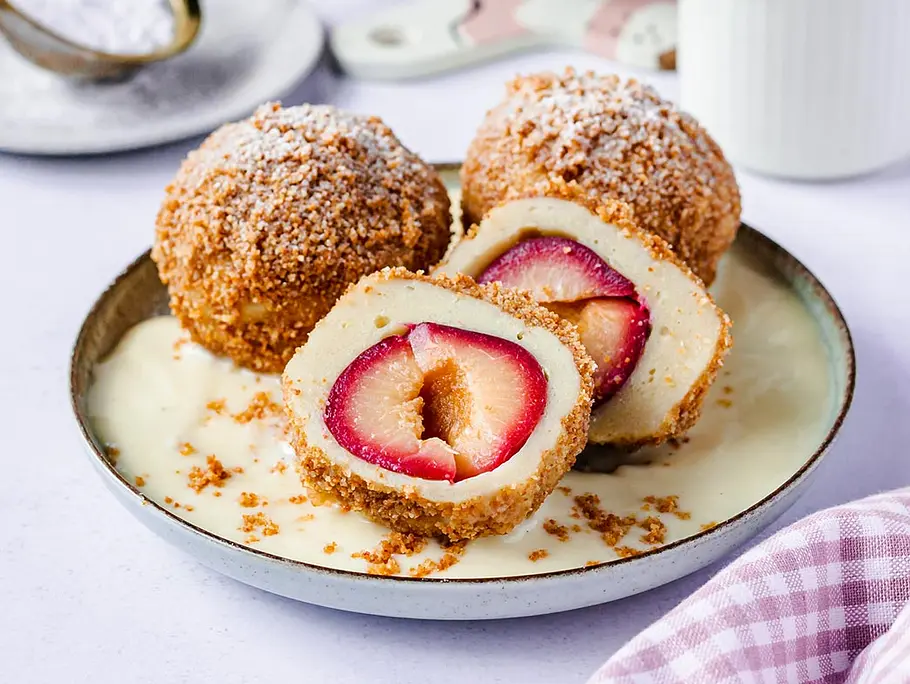 Drei Zwetschgenknödel sind auf einem Teller mit Vanillesoße serviert. Zu erkennen ist die knusprige Bröselkruste. Ein Pflaumenknödel ist halbiert, sodass man die Frucht erkennt.