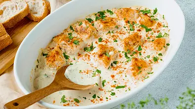 Philadelphia-Hähnchen in einer weißen Auflauflaufform in der ein Holzlöffel liegt. Ein Stück Hähnchen wurde rausgenommen. - Foto: ShowHeroes für LECKER @ Bauer Media Group