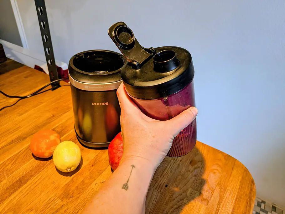 Eine Hand hält den gefüllten Mixbehälter des Philips 5000 Series Smoothiemaker, daneben stehen Gerät und Obst auf der Holzplatte.
