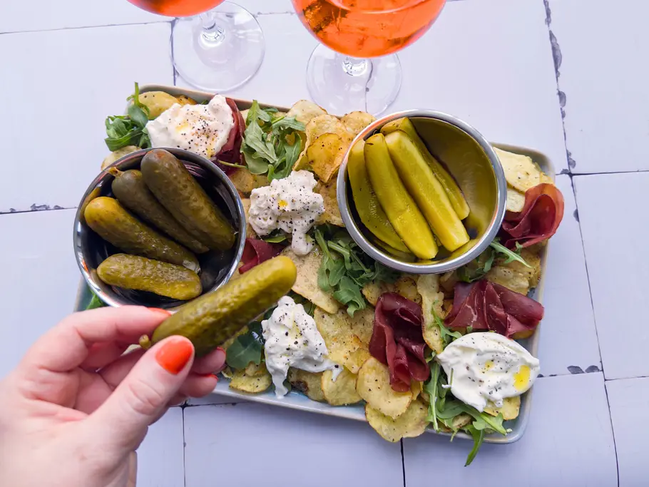 Topshot auf ein rechteckiges Pickle Board, auf dem Chips mit einem Belag aus Bresaola-Rosen, zerzupftem Burrate, Rucola und Gewürzgurken in Edelstahlschüsseln liegen, nach denen eine Hand greift.