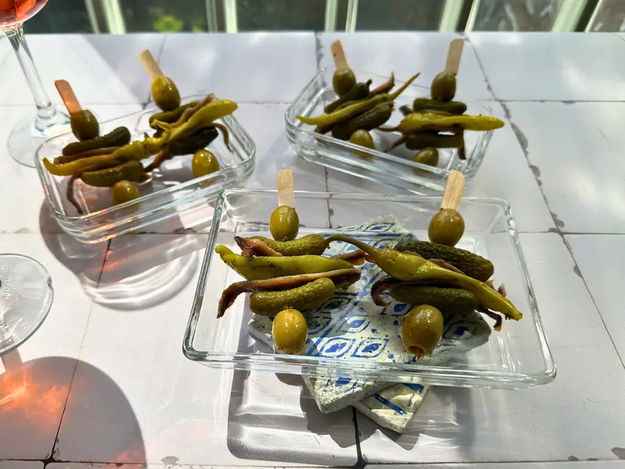 Auf einer hellen Fliesenfläche stehen drei eckige Glasschalen mit je zwei Partyspießen, auf denen grüne Oliven, Anchovi und Cornichons aufgereiht sind.