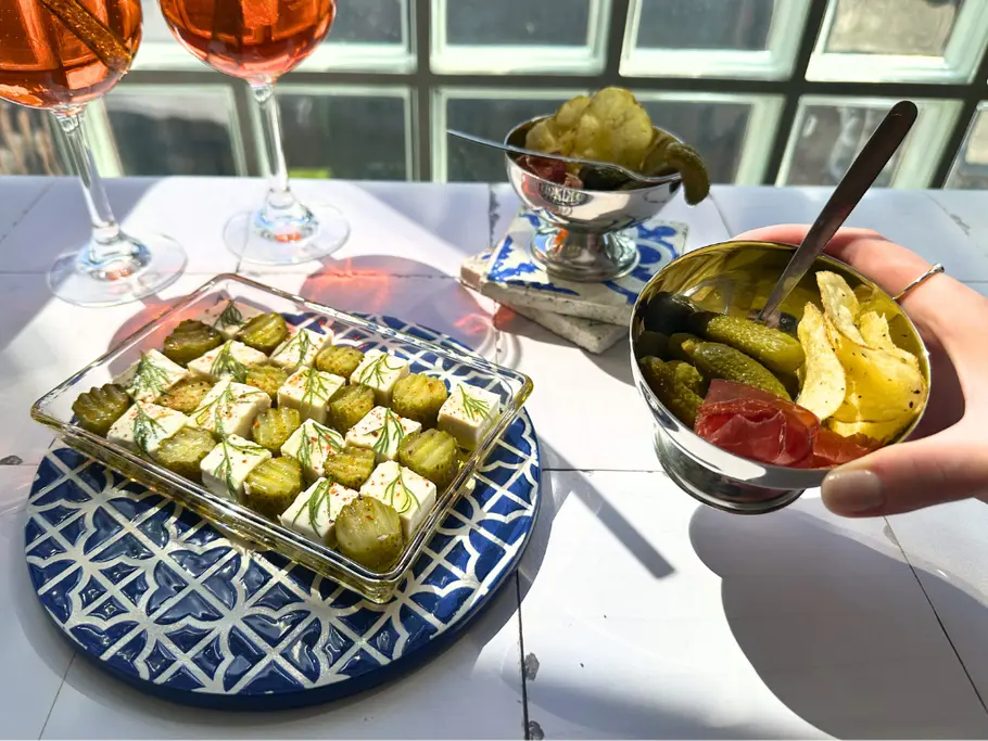Auf einem Tisch steht eine eckige Glasschale mit schachbrettartig angeordneten Fetawürfeln und sauren Gurkenstücken, die mit Olivenöl beträufelt und mit Dill und Chiliflocken garniert sind, neben einer Edelstahl-Eisschale mit Chips, Cornichons und Bresaola.