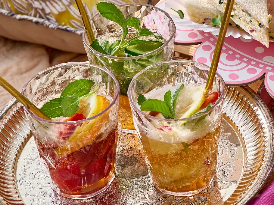Drei Gläser Pimm’s Cup mit Eiswürfeln, Zitronen- und Limettenscheiben sowie frischer Minze auf einem silbernen Tablett serviert.