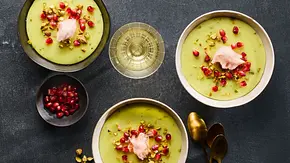 Drei Schalen mit cremigem, grünem Pistazien-Pudding, garniert mit Granatapfelkernen, gehackten Pistazien und einem Klecks rosa Zuckerwatte auf dunkler Unterlage. Daneben ein Schälchen mit weiteren Granatapfelkernen und goldene Löffel. - Foto: LECKER @ Bauer Media Group