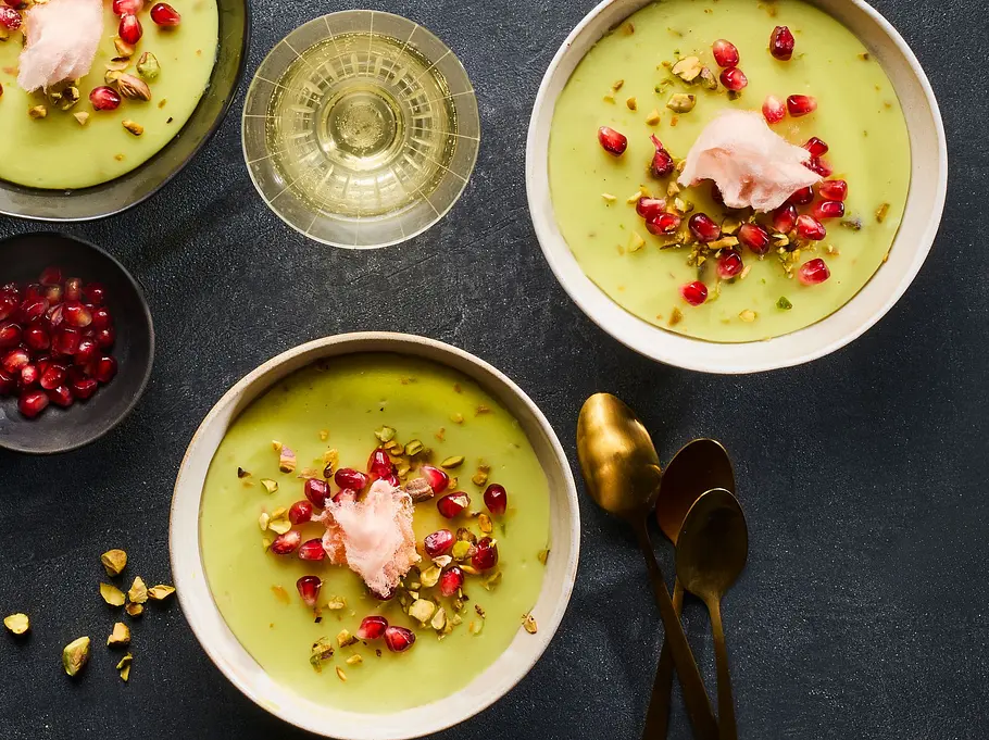 Drei Schalen mit cremigem, grünem Pistazien-Pudding, garniert mit Granatapfelkernen, gehackten Pistazien und einem Klecks rosa Zuckerwatte auf dunkler Unterlage. Daneben ein Schälchen mit weiteren Granatapfelkernen und goldene Löffel.