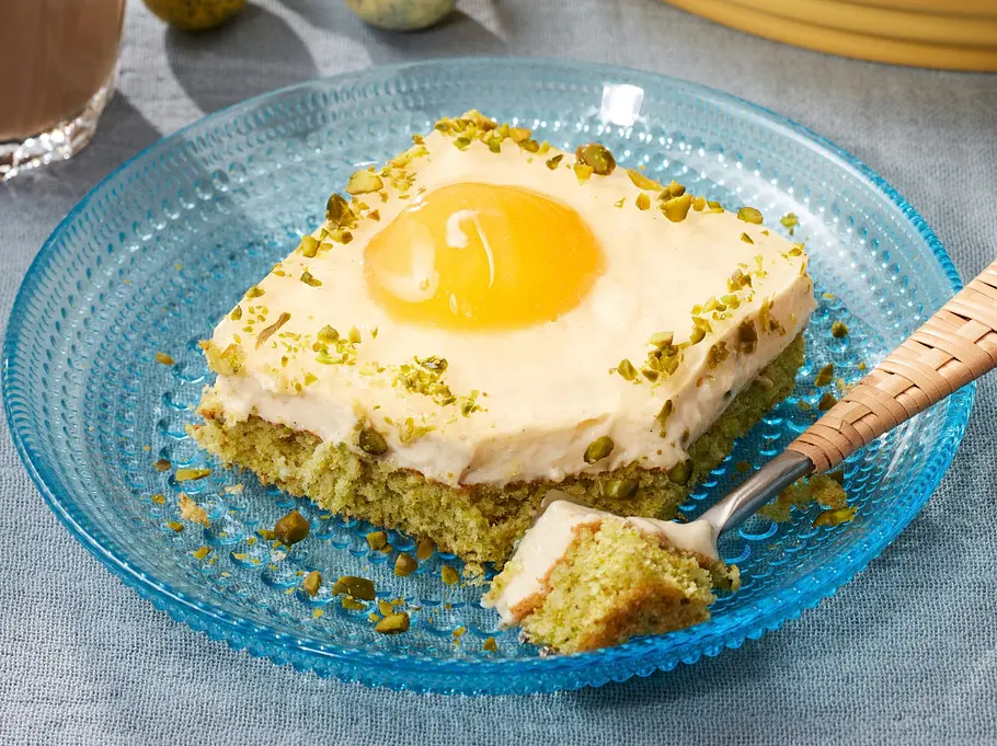 Pistazien-Spiegelei-Kuchen in eckigen Stücken auf einer gelben Platte und einem blauen Teller angerichtet, mit grünem, lockerem Pistazienboden, weißer Creme und einem fruchtigen Aprikosen-Topping, das wie ein Spiegelei aussieht, bestreut mit gehackten Pistazien.