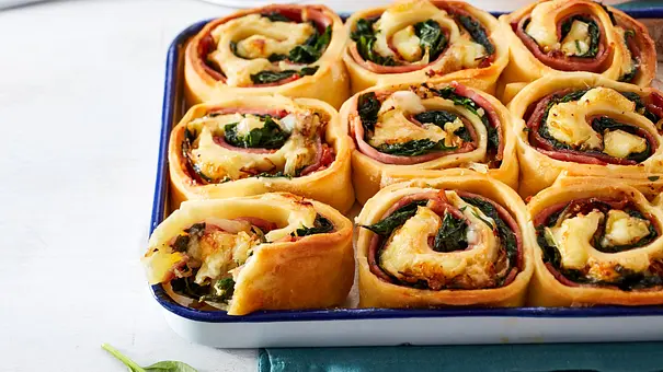 Goldbraune Pizza-Schnecken mit geschmolzenem Käse, Kasseler und frischem Spinat in einer weißen Auflaufform, angerichtet auf heller Holzoberfläche. - Foto: LECKER @ Bauer Media Group