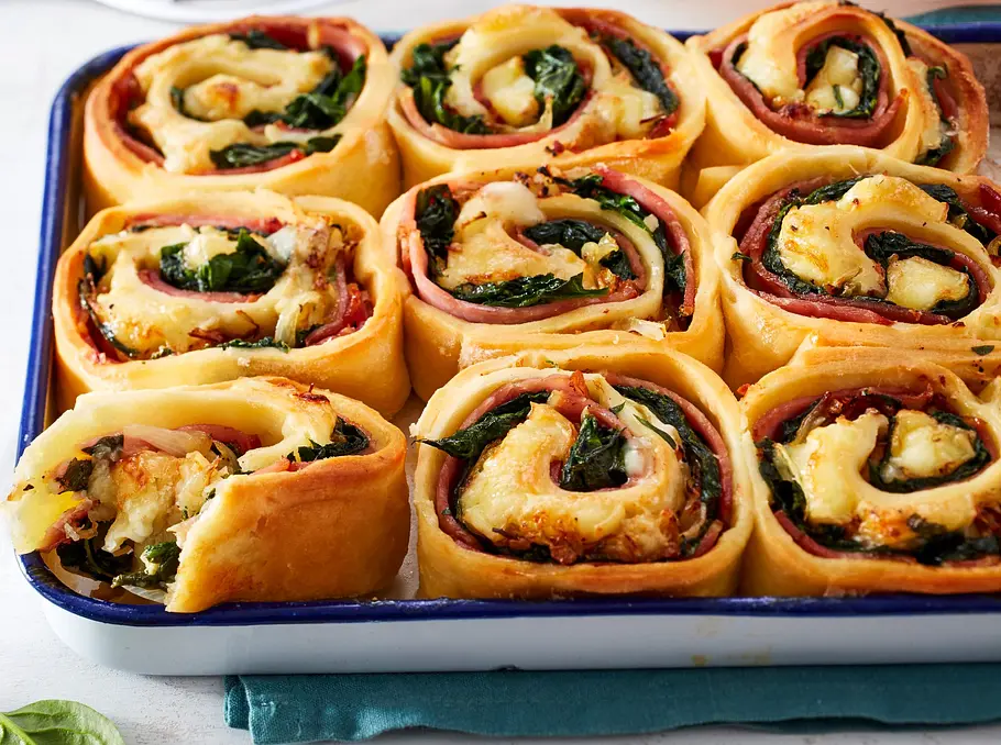 Goldbraune Pizza-Schnecken mit geschmolzenem Käse, Kasseler und frischem Spinat in einer weißen Auflaufform, angerichtet auf heller Holzoberfläche.