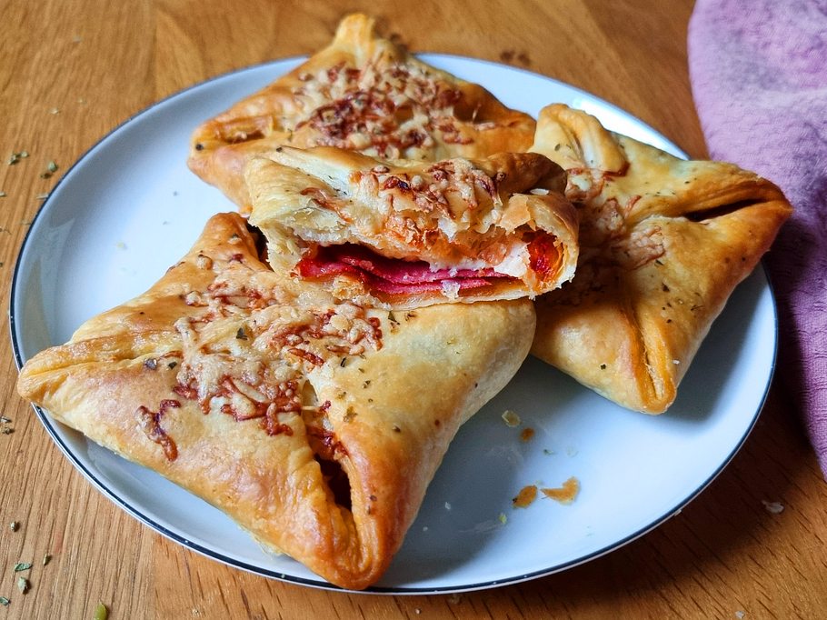 Die Blätterteig-Pizzataschen liegen auf einem Teller. Eine Pizzatasche ist angeschnitten, sodass man die Füllung aus Salami und Tomatensoße erkennt.