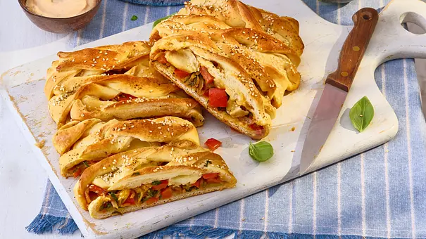 Aufgeschnittener, goldbrauner Pizza-Strudel mit Gemüse- und Käsefüllung, serviert auf einem weißen Schneidebrett mit Sesam bestreut, daneben ein Messer und Dip. - Foto: LECKER @ Bauer Media Group