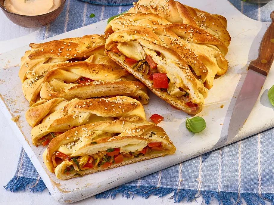 Aufgeschnittener, goldbrauner Pizza-Strudel mit Gemüse- und Käsefüllung, serviert auf einem weißen Schneidebrett mit Sesam bestreut, daneben ein Messer und Dip.