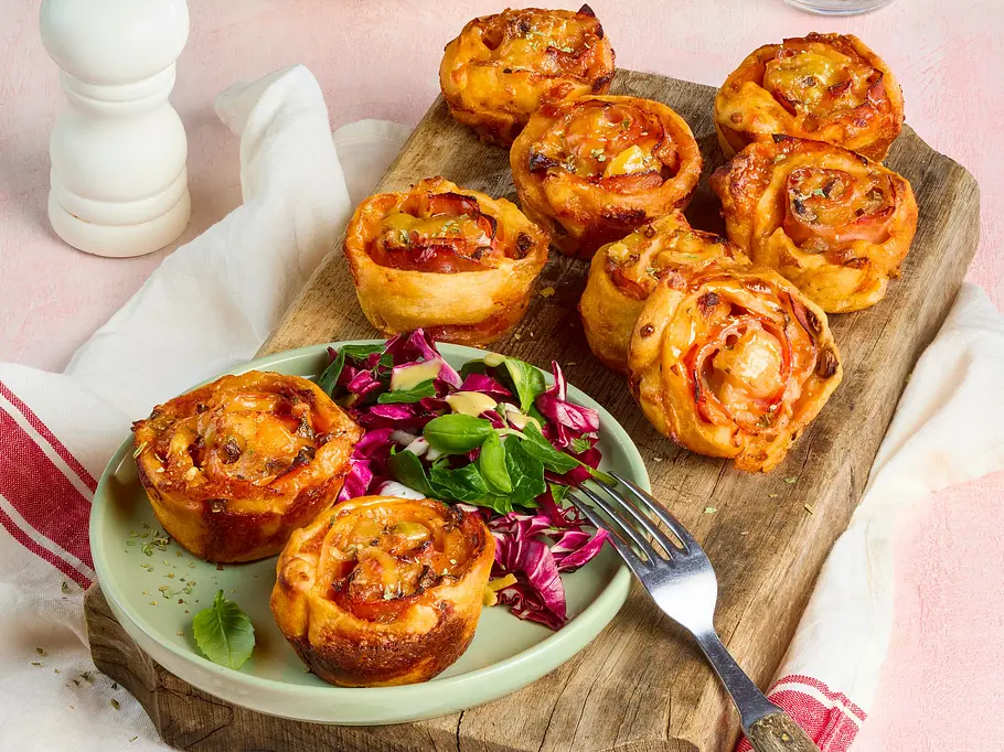 Pizza-Muffinschnecken Goldbraune Teigschnecken liegen auf einem Holzbrett und einem grünen Teller mit Blattsalat. Dahinter stehen Salat und ein Glas.