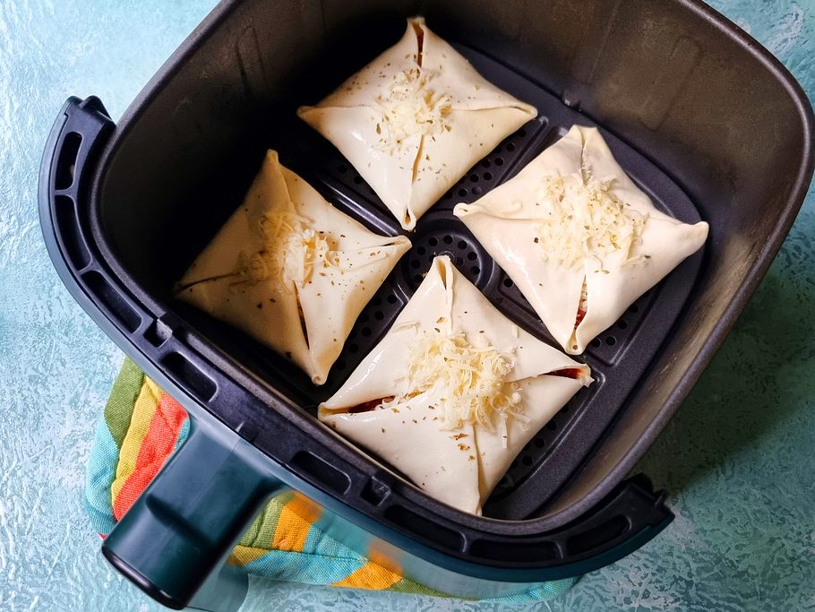 4 ungebackene Pizzataschen liegen im Garkorb der Heißluftfritteuse und sind mit ein wenig Käse bestreut.