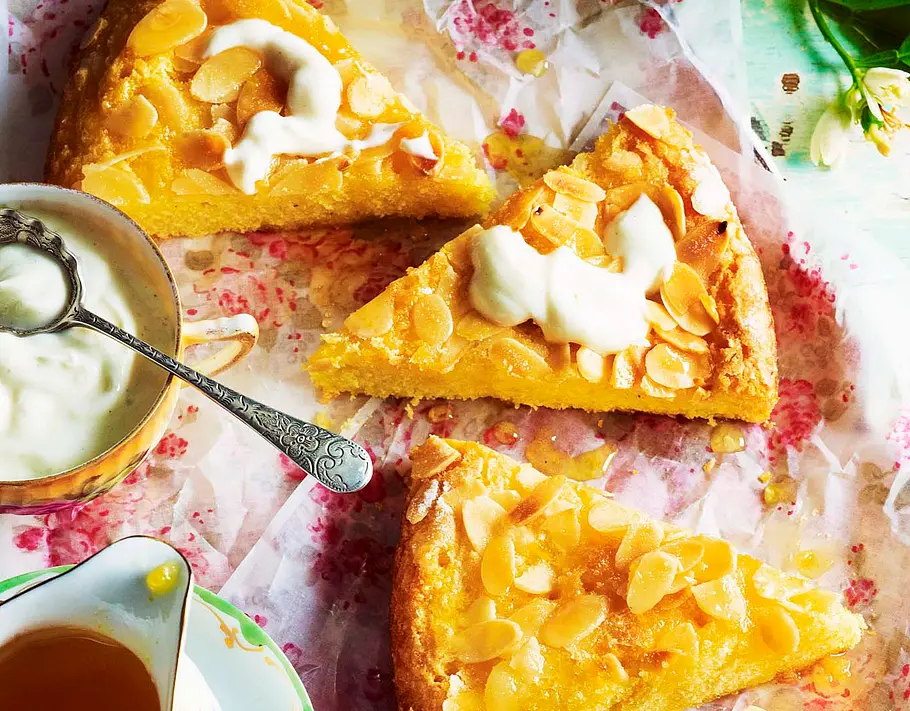 Polenta-Orangen-kuchen mit Mandeln Rezept Drei Stücke Polenta-Orangen-Kuchen mit Mandeln und Joghurttupfen liegen auf buntem Backpapier, daneben stehen Joghurt und Sirup.