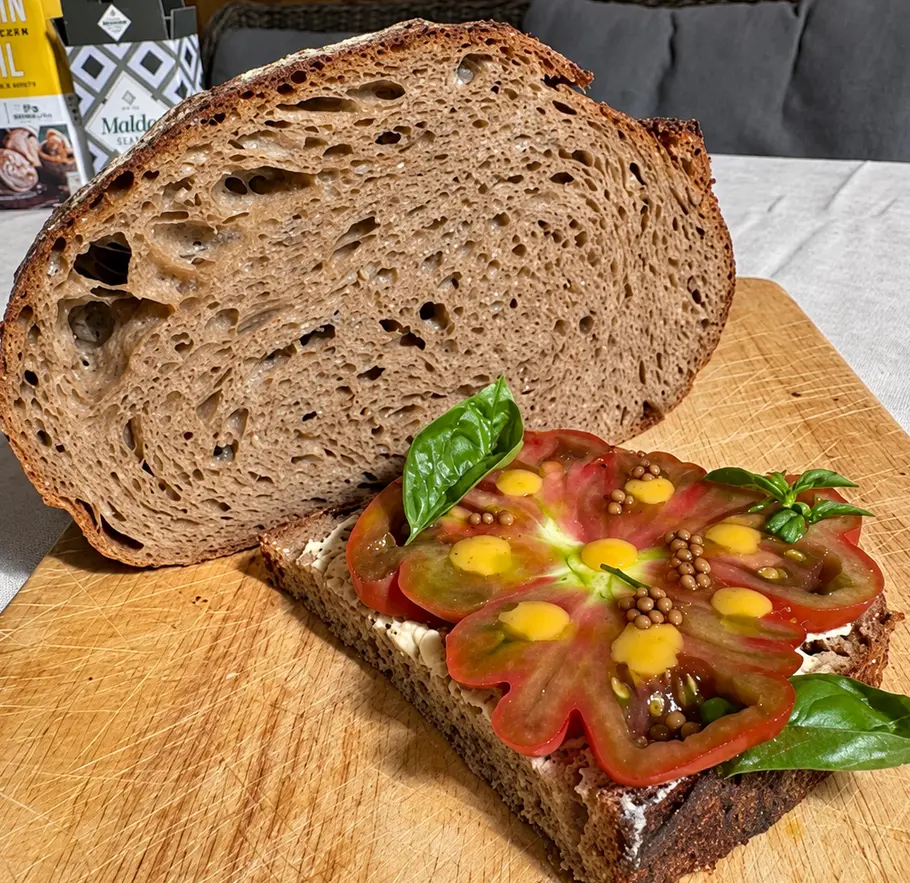 Protein-Sauerteigbrot mit Tomate Auf einem Holzbrett liegen ein angeschnittenes Brot und eine Scheibe mit Tomatenscheiben, gelben Tupfen und Basilikum.
