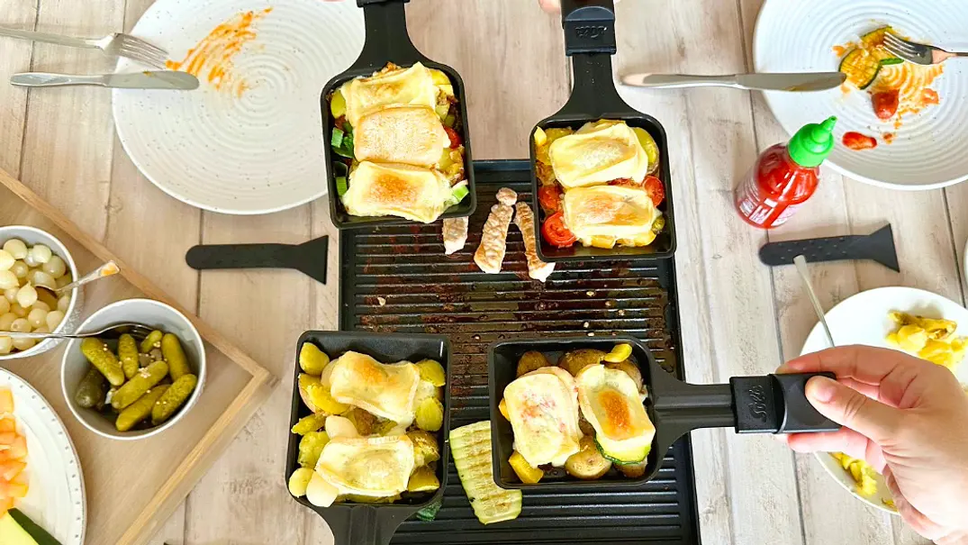 Topshot auf einen Raclette-Grill, über den vier Hände ihre Pfännchen mit geschmolzenem und gebräuntem Raclette-Käse halten - Foto: LECKER @ Bauer Media Group / Nicole Molitor