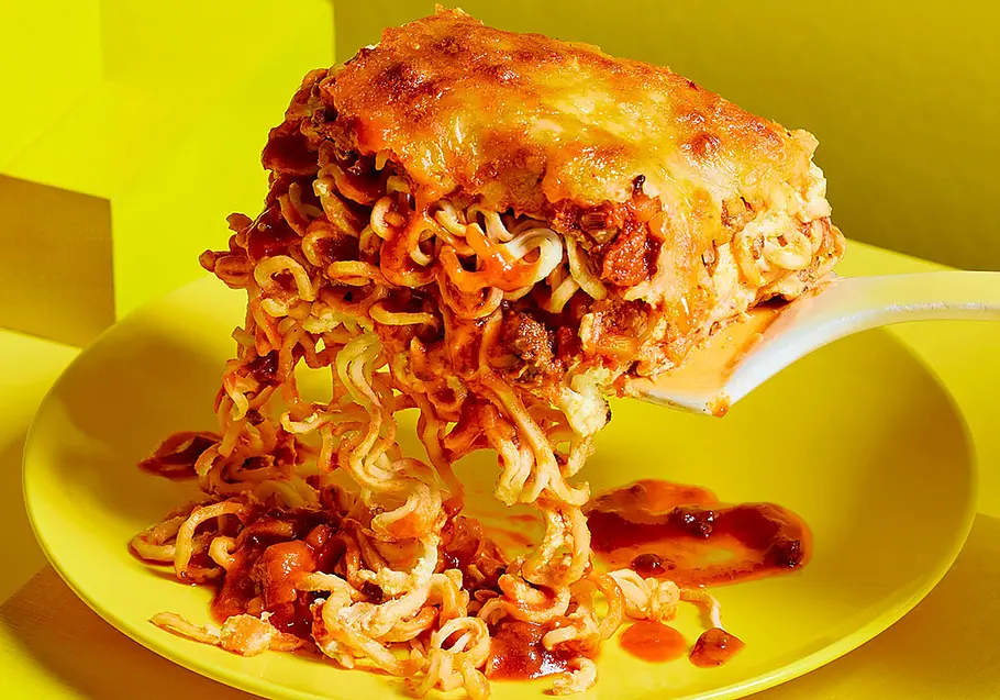 Ein weißer Pfannenwender hebt eine Portion käseüberbackene Nudeln mit roter Sauce über einen gelben Teller.