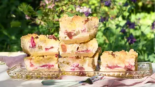 Saftige Stücke Rhabarber Crumble Cake mit goldenen Streuseln und zarten rosa Rhabarberstückchen im hellen Teig, locker gestapelt auf einer länglichen Glasplatte. Daneben liegen kleine Gabeln auf einer Stoffserviette, die Kuchenstücke wirken weich und fruchtig mit knuspriger Oberfläche. - Foto: LECKER @ Bauer Media Group