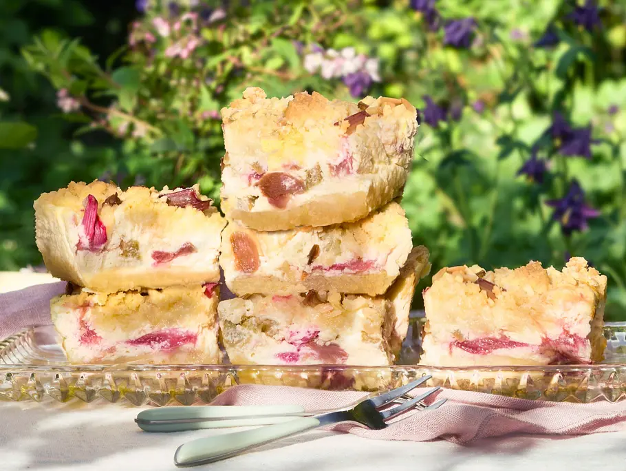 Saftige Stücke Rhabarber Crumble Cake mit goldenen Streuseln und zarten rosa Rhabarberstückchen im hellen Teig, locker gestapelt auf einer länglichen Glasplatte. Daneben liegen kleine Gabeln auf einer Stoffserviette, die Kuchenstücke wirken weich und fruchtig mit knuspriger Oberfläche.