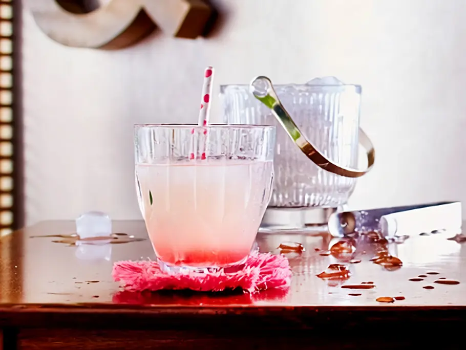 Ein Glas Rhabarber-Limonade mit Papierstrohhalm auf einem pinken Stoff-Untersetzer im Hintergrund ein Eiswürfelbehälter aus Glas.