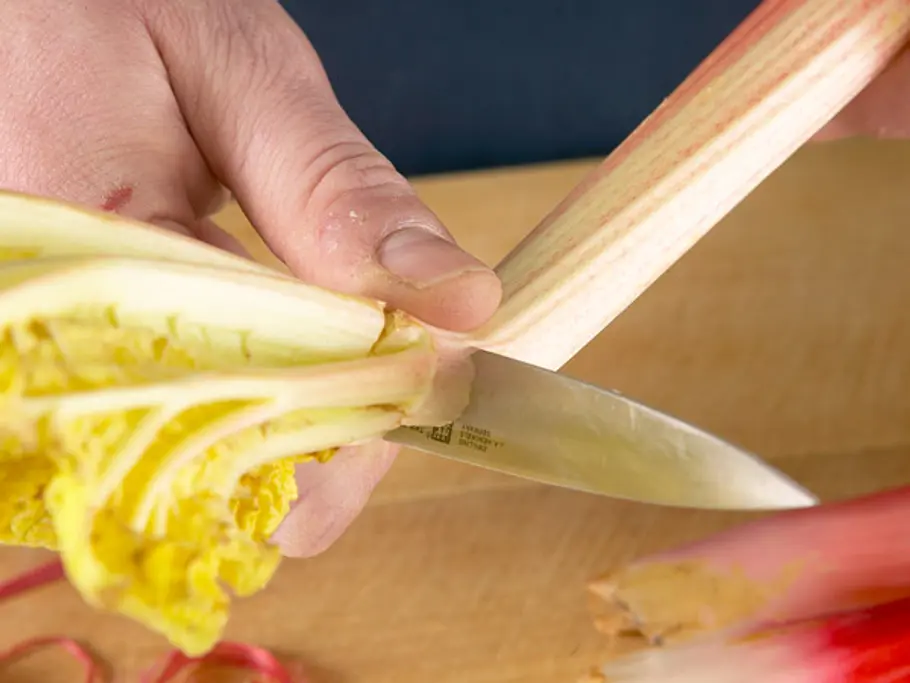 Mit einem Messer wird das Blatt und Wurzelende von einer frischen Rhabarberstange abgeschnitten, darunter ein Holzbrett.
