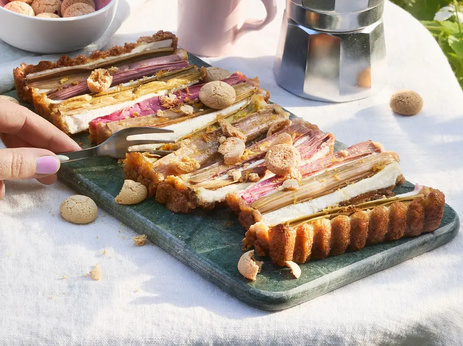 Rhabarber-Tarte mit knusprigem, goldbraunem Rand und einer Füllung aus hellen Creme- und rosa Rhabarberstreifen, in Stücke geschnitten und mit zerbröselten Amarettini bestreut. Serviert auf einer dunklen Platte, daneben eine Schale mit Amarettini und eine Tasse Kaffee auf einer hellen Tischdecke.