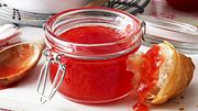 Rhabarbermarmelade Rezept: Ein Bügelglas ist mit der roten Konfitüre gefüllt. Daneben liegt ein Stück Croissant, von dem etwas der Rhabarbermarmelade herunterläuft. Im Hintergrund ist ein Kochlöffel und ein -topf zu erkennen.  - Foto: House of Food / Bauer Food Experts KG