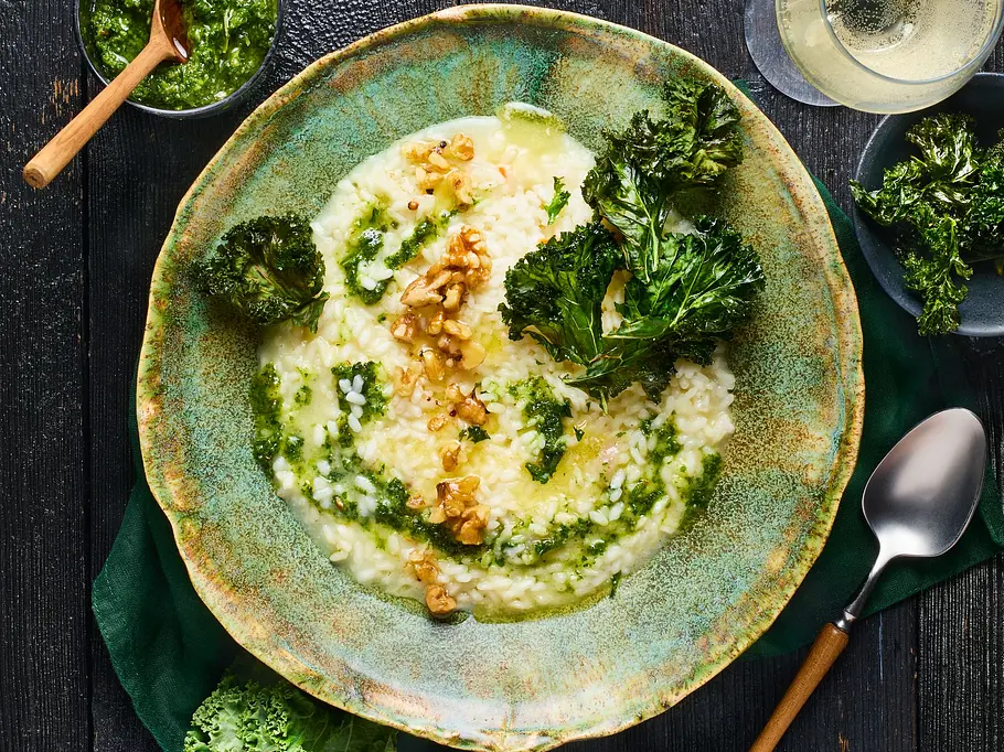 Cremiges Risotto, serviert in einer rustikal-grünen Schale, getoppt mit grünem Grünkohl-Pesto, knusprigen Grünkohlchips und Walnüssen, daneben kleiner Teller mit Grünkohlchips und ein Glas Weißwein.