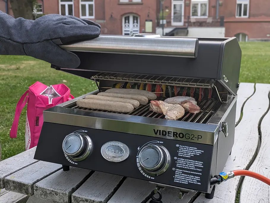 Rösle Videro G2-P Grillhandschuh Hand mit Grillhandschuh hebt den Deckel des Videro G2-P, auf dem Grill liegen Würstchen und Hähnchenteile.