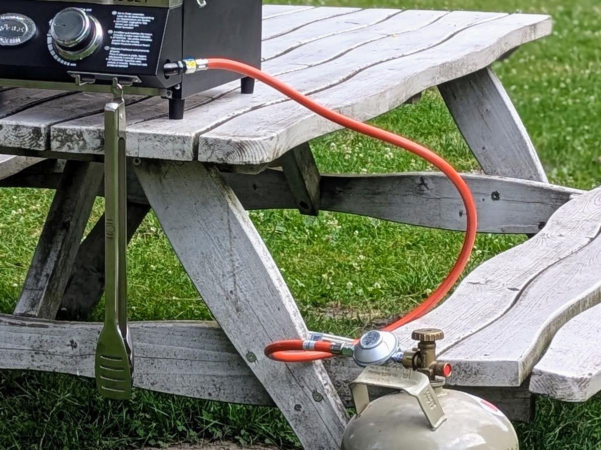 Rösle Videro G2-P Tischgrill Camping-Gasgrill Videro G2-P mit angeschlossener Gasflasche steht geschlossen auf einer Picknickbank im Freien.