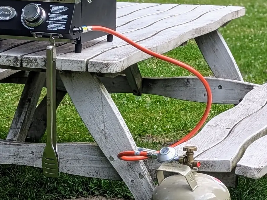 Rösle Videro G2-P Tischgrill Camping-Gasgrill Videro G2-P mit angeschlossener Gasflasche steht geschlossen auf einer Picknickbank im Freien.