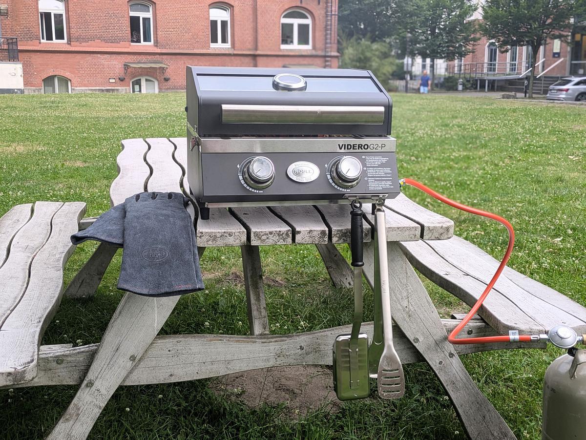 Rösle Videro G2-P Zubehör Videro G2-P Grill auf Picknickbank mit Grillhandschuhen, Grillzange und Grillbürste.