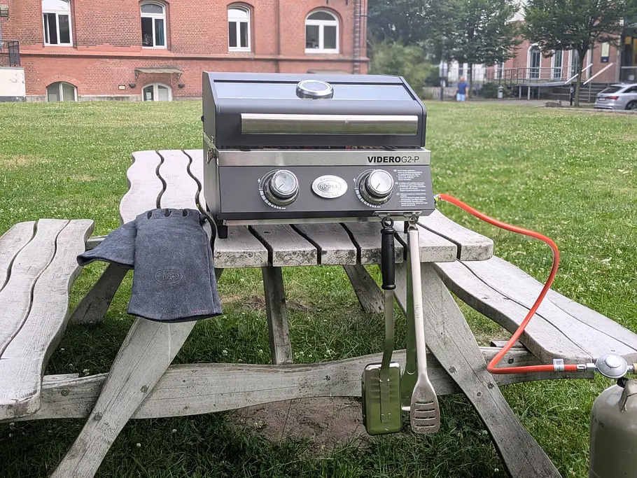 Rösle Videro G2-P Zubehör Videro G2-P Grill auf Picknickbank mit Grillhandschuhen, Grillzange und Grillbürste.