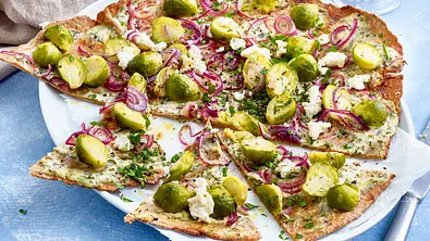 Auf einem weißen Teller liegt knuspriger Flammkuchen mit Rosenkohl, roten Zwiebelringen, weißen Käsebröckchen und Kräutern. - Foto: LECKER @ Bauer Media Group Auf einem weißen Teller liegt knuspriger Flammkuchen mit Rosenkohl, roten Zwiebelringen, weißen Käsebröckchen und Kräutern. - Foto: LECKER @ Bauer Media Group
