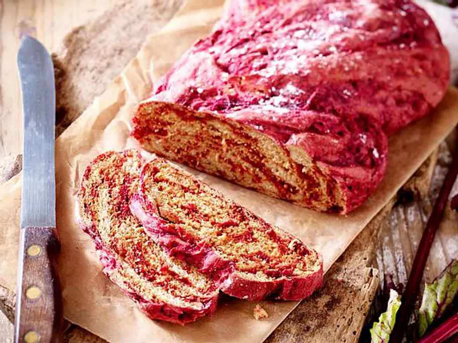 Rote-Bete-Hefezopf auf Backpapier, angeschnitten mit sichtbarer rosa Marmorierung und zwei Scheiben auf rustikalem Holzbrett.