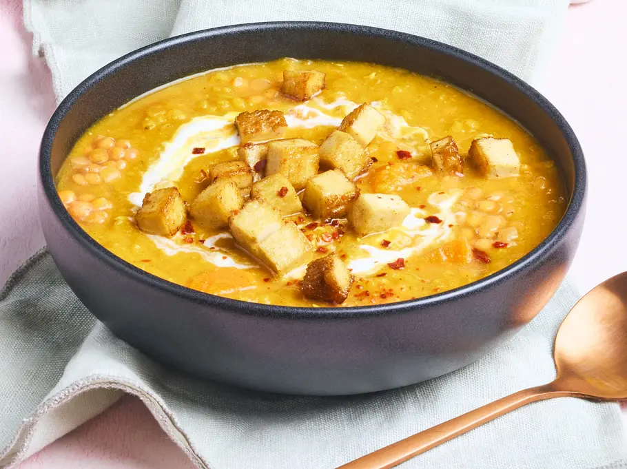 Rotes Linsen-Dal in einer Schale, garniert mit knusprigen Tofu-Croûtons und einem Klecks Joghurt, dazu etwas Chili und serviert auf einem hellen Tisch mit Getränk und Blumen im Hintergrund.