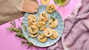 Runde, goldene Kräuter-Cookies mit frischen Kräuterblättchen auf einem hellblauen Teller, daneben Zweige von Rosmarin und Thymian sowie ein Glas mit Kräutern auf einem rosa Untergrund. - Foto: LECKER @ Bauer Media Group Runde, goldene Kräuter-Cookies mit frischen Kräuterblättchen auf einem hellblauen Teller, daneben Zweige von Rosmarin und Thymian sowie ein Glas mit Kräutern auf einem rosa Untergrund. - Foto: LECKER @ Bauer Media Group