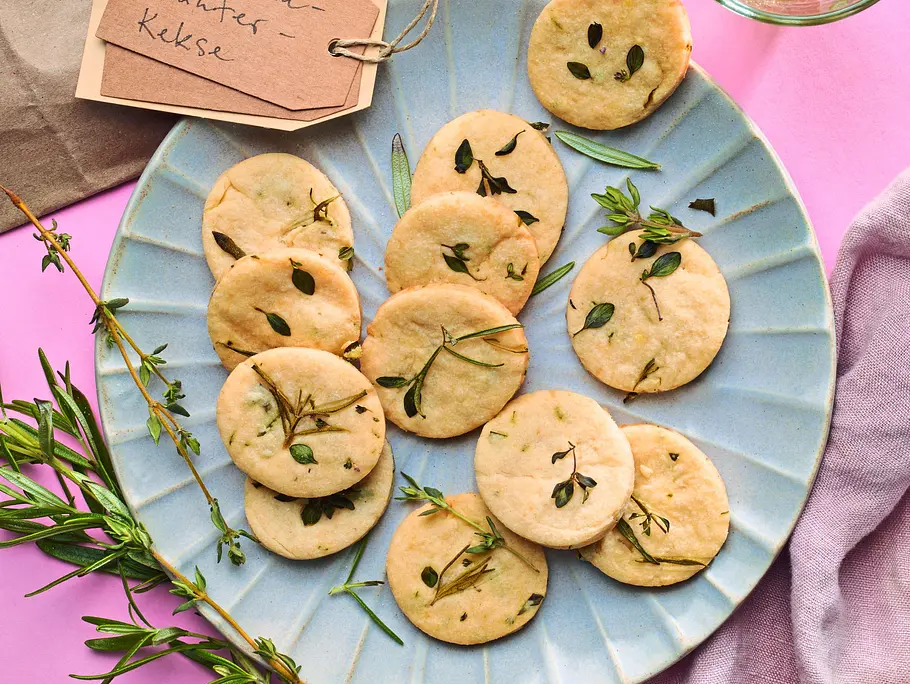 Runde, goldene Kräuter-Cookies mit frischen Kräuterblättchen auf einem hellblauen Teller, daneben Zweige von Rosmarin und Thymian sowie ein Glas mit Kräutern auf einem rosa Untergrund.