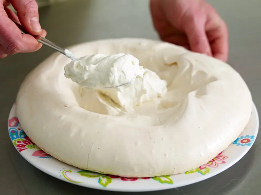 Steif geschlagene Sahne wird mit einem Löffel in die Mulde einer gebackenen Pavlova auf einem bunten Teller gefüllt.