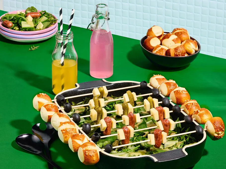 Salat in einer Auflaufform, darauf Laugenspieße mit Mini-Laugenbrötchen, Käsewürfeln, Oliven, Würstchen und Cornichons.